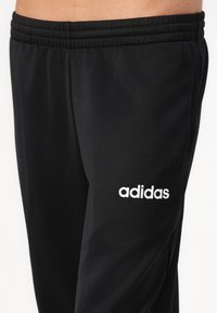 Fekete sportnadrág elasztikus derékkal és háromsoros varrással. Az alsó lábszárán egy fehér "adidas" logó található. Simaszerkezet.