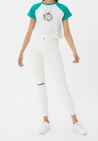 Bílý zkrácený tričko s tyrkysovými raglánovými rukávy, s grafikou vintage televize a textem "peace". V páru s bílými skinny džínami.