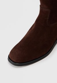 Stivale alla caviglia in suede marrone con una texture liscia, punta arrotondata e suola in gomma nera. Presenta un design semplice con dettagli di cucitura minimi.