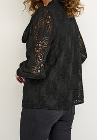 Kvinna klädd i en svart spetsblus med blommönster och puffärmar, i kombination med mörka, figursydda jeans, vänd sidledes mot en neutral bakgrund.