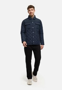 Giacca trapuntata blu navy con due tasche frontali, bottoni a pressione e colletto. Pantaloni neri e sneaker grigie e bianche completano l'outfit.