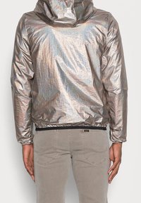 Metallisch zilver hooded jack met een gestructureerde afwerking. Voorzien van elastische manchetten en zoom. Draag het over lichtgrijze jeans.