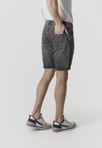 Mann steht mit einer Hand in der Tasche seiner grauen Jeansshorts, trägt weiße Sneakers und ein weißes T-Shirt vor einem einfarbigen Hintergrund.