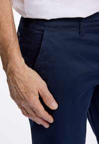 Pantaloni in misto cotone blu navy con una superficie liscia, dotati di tasche laterali e una cintura sicura, con una mano poggiata sulla gamba.