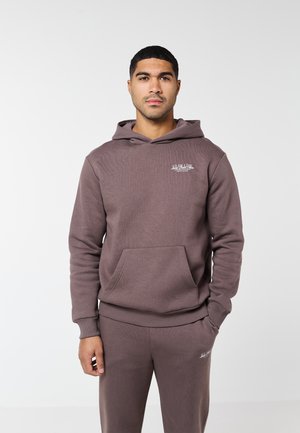 Férfi áll szemben, mauve színű, egyező kapucnis pulóvert és melegítőnadrágot viselve, fehér feliratokkal a mellkason és a comb felső részén.