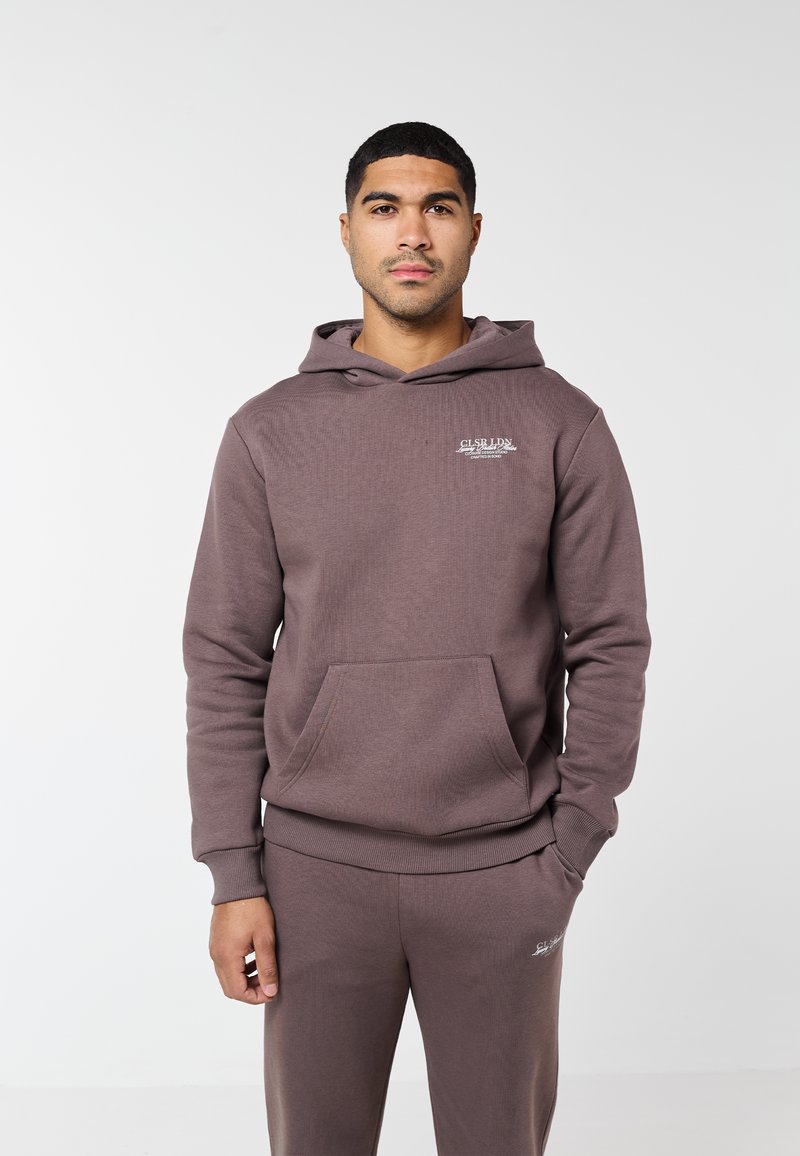 Férfi áll szemben, mauve színű, egyező kapucnis pulóvert és melegítőnadrágot viselve, fehér feliratokkal a mellkason és a comb felső részén.