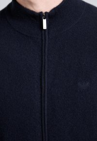 Detailný záber na tmavomodrú zipsovú mikinu s rebrovaným golierom a jemne vyšitým logom na hrudi.