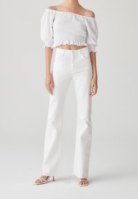 Haut blanc court à épaules dénudées avec smocks texturés, associé à un jean blanc évasé taille haute et des talons beiges à brides.