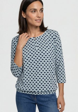 Vrouw draagt een lichtblauwe blouse met donkerblauw cirkelpatroon en driekwart mouwen, staand tegen een effen achtergrond.