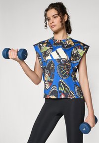 Haut sans manches à motif de papillons bleus, avec des accents aux épaules et un logo blanc, accompagné de leggings noirs et tenant un haltère bleu.