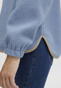 Ljusblå fleeceöverdel med mjuk textur, med elastiska ärmslut och rundad nederkant med beige detaljer. Bärs över mörk denim.