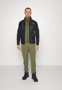 Gilet isolato nero con texture trapuntata, base layer verde oliva, pantaloni abbinati e scarponi da trekking neri, posizionati su uno sfondo semplice.
