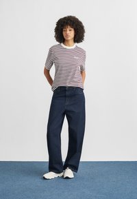 Γυναίκα με σγουρά μαλλιά στέκεται σε μπλε χαλί φορώντας λευκό και μωβ ριγέ πουκάμισο, φαρδιά σκούρα τζιν και λευκά αθλητικά παπούτσια.