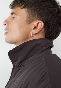 Schwarze Jacke mit hohem Kragen und glatter Textur, mit sichtbaren Nähten und einem runden Kragen, getragen von einer Person mit einer geschwungenen Silhouette.