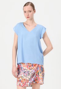 Camicia azzurro chiaro con scollo a V e maniche corte, abbinata a una gonna colorata e patterned con vortici di tonalità arancione, rosa e blu.