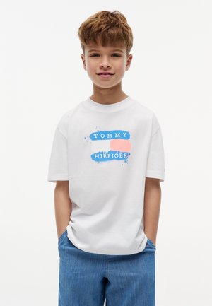 Dreng med kort brunt hår smiler, iført en hvid Tommy Hilfiger t-shirt og blå denimshorts, hænderne i lommerne.