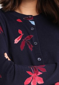 Mörkblå kofta med blommönster i röd och rosa, med rund halsringning, sex knappar och långa ärmar. Mjuk stickad textur.