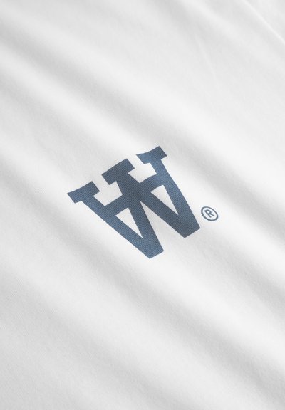 Weißes T-Shirt aus Stoff mit einem großen, dunkelblauen grafischen Logo und einem eingetragenen Markenzeichen-Symbol. Die Textur erscheint glatt und weich.