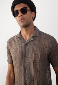 Cămașă tricotată maro cu mânecă scurtă, având un model texturat și două butoane, purtată cu ochelari de soare negri și stilizată pentru un look modern.