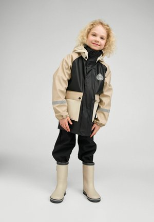 Małe dziecko z kręconymi blond włosami, ubrane w beżowo-czarną kurtkę przeciwdeszczową, czarne spodnie i beżowe kalosze, stojące na jednolitym tle.