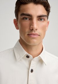 Lichtbeige overhemd met knopen, voorzien van een kraag, twee bruine knopen en een gestructureerde stof. Close-up van het shirt en de halslijn.