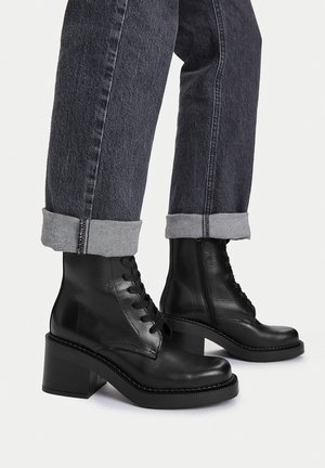 Beine, die dunkelgraue, hochgekrempelte Jeans und schwarze Leder-Stiefeletten mit Schnürung und Blockabsatz auf weißem Hintergrund tragen.