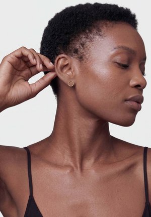 Közeli kép egy rövid, göndör hajú nőről, aki egy kis aranykarika fülbevalót visel, megérinti a fülét, és oldalra néz csukott szemmel.