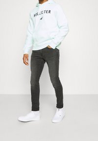 Ljus mintgrön hoodie med "HOLLISTER"-logotyp, framficka, grå slim-fit jeans och vita sneakers. Enkelt mönster och mjuka tyger.