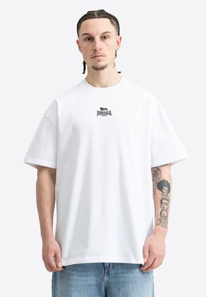Man met gevlochten haar die een wit Lonsdale London T-shirt en blauwe spijkerbroek draagt, toont zijn getatoeëerde linker onderarm met zwarte inktontwerpen.
