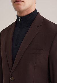 Hombre vistiendo un blazer marrón oscuro sobre un suéter negro con cremallera y cuello acanalado, mostrando la parte superior del pecho y el área del cuello.