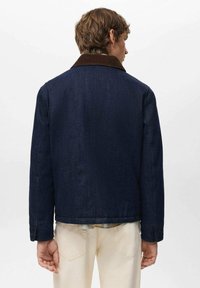 Temno modra denim jakna z rjavim kordurovnim ovratnikom, ravnim krojem in gumbi na zapestjih. Vidna od zadaj, oblečena preko karo srajce.