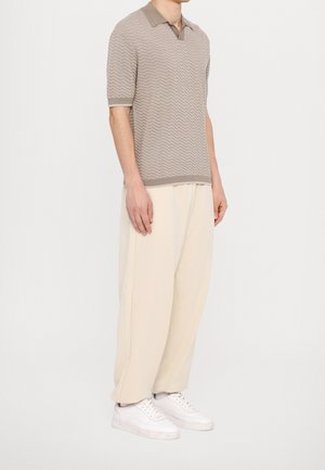 Homme portant un polo en maille beige à manches courtes avec motif, un pantalon ample crème et des baskets blanches, debout devant un fond blanc uni.