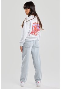 Light gray sweatshirt with graphic design on back, paired with light blue baggy jeans. Model wears white sneakers with red accents.