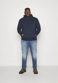 Tamsiai mėlynas džemperis su kapišonu ir priekyje kišene bei raišteliais, derinamas su šviesiai mėlynais plyšytais džinsais. Modelis dėvi alyvuogių žalią batus.