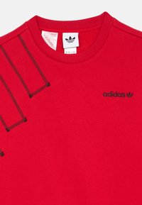 Červená bavlnená mikina s rebrovaným okrúhlym výstrihom. Obsahuje čierne akcenty a malú čiernu značku Adidas na hrudníku.