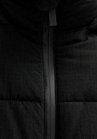 Chaqueta de plumas negra con textura acolchada, detalles de costuras verticales y cierre frontal con cremallera de acabado mate.