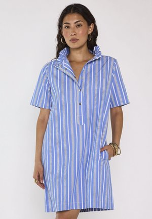 Femme portant une robe à manches courtes rayée verticalement bleue et blanche avec un col montant à volants et la main dans une poche.
