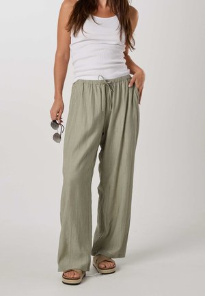 Femme portant un pantalon ample vert clair, un top blanc sans manches, des sandales beiges, tenant des lunettes de soleil dans une main et l’autre main dans la poche.