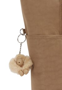 Tan fabric bag with a side pocket, featuring a fluffy beige monkey keychain attached by a metal ring. Keychain has small limbs and facial detail.