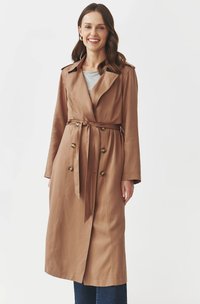 Ruskea trench-coat, jossa on vyöllinen vyötärö, kaksoisnapitus ja hartiapolttimet. Kangas on sileä, yhdistetty harmaaseen toppiin ja tummaan farkkuun.