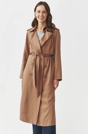 Hnědý trench coat s páskem v pase, dvouřadým designem a ramenními epauletami. Tkanina má hladkou texturu, v kombinaci se šedým topem a tmavými džíny.