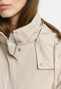 Beige vandtæt jakke med høj krave, lynlås lukning og trykknapper. Glat stof med minimalistisk design og snor detaljer.