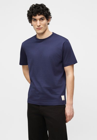 Άνδρας με σγουρά μαλλιά που φοράει μπλε κοντομάνικη μπλούζα και μαύρο παντελόνι, στέκεται μπροστά σε μονόχρωμο ανοιχτό φόντο.