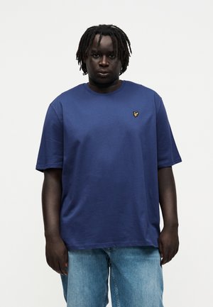 Personne avec des dreadlocks courtes portant un t-shirt bleu avec un petit logo d'oiseau jaune et un jean bleu clair, debout devant un fond blanc uni.