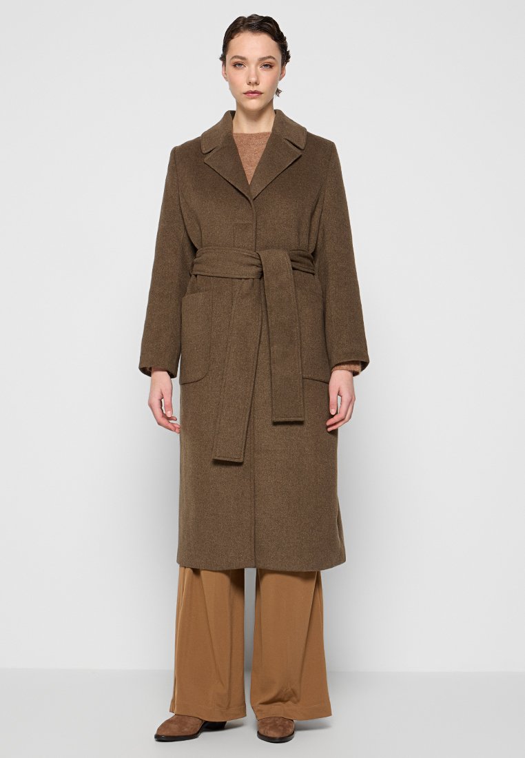 Brauner, langer Wollmantel mit Reverskragen, tailliert mit Gürtel und zwei Fronttaschen. Getragen über beigen Weitbein-Hosen und braunen Schuhen.