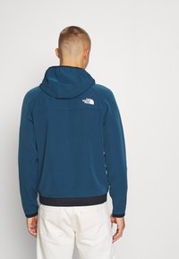 Kurtka z kapturem w ciemnym kolorze teal, z dopasowanym krojem, elastycznymi mankietami i prążkowanym dołem. Logo w białym kolorze na górnej części pleców.