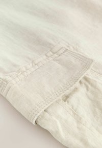 Tela de lino de color beige claro con un puño doblado, mostrando detalles de costura visibles. La textura parece suave y ligeramente arrugada.