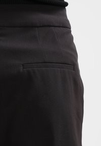 Pantalon noir sur mesure avec une texture lisse, doté d'une poche passepoilée unique et de plis subtils pour un look structuré.