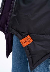 Černá péřová bunda s prošívanou texturou, s špičatým lemem. Oranžová visačka s nápisem "VIDĚT VŠE" a grafikou slunce. Viditelný detail zipu.