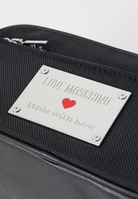Čierna textúrovaná taška so striebornou logom doskou; vygravírované "LOVE MOSCHINO" a "Vyrobené s láskou," doplnené červeným srdcom. Viditeľný detail zipsu.
